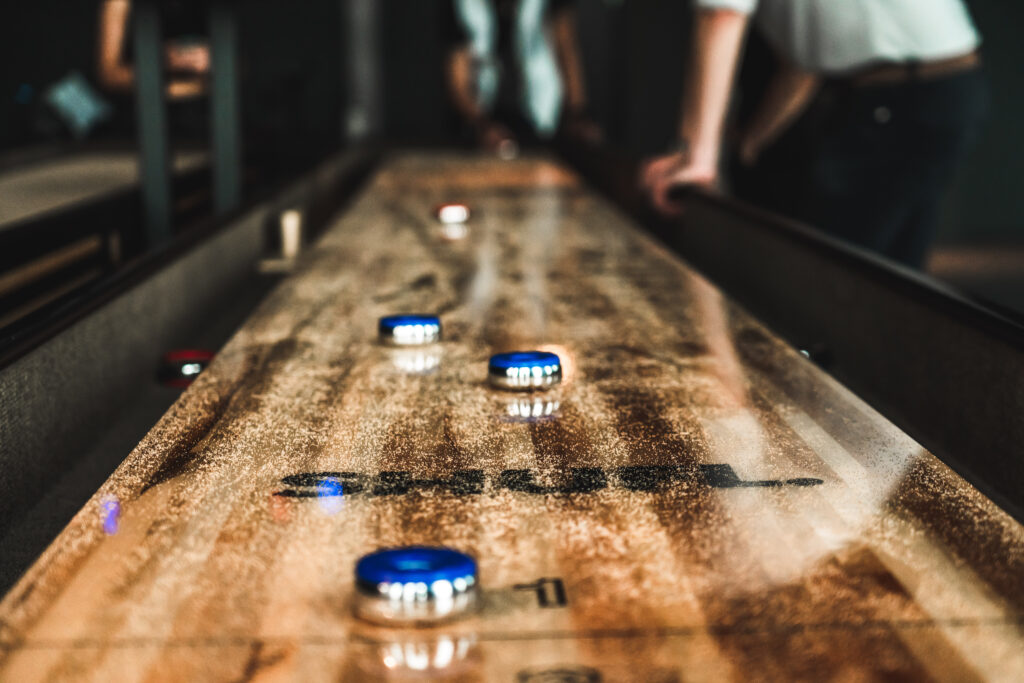 Et shuffle board midt i et spill, en person står på enden og er klar til å spille, og en person lener seg på bordet.